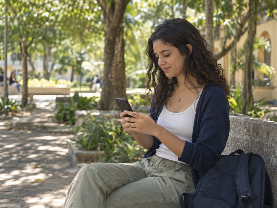 Estudante sentado num banco ao ar livre, procurando kitnet pelo celular.